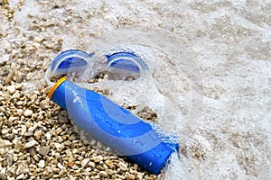 Swimming goggles and suntan lotion lying on pebble beach in water.