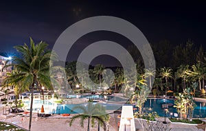 Swimming pool area in a beach hotel resort during night with trees and smooth blue swimming pool water reflecting lights long