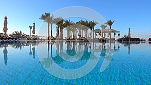 Swimming pool with clear blue water, palm trees, deck chairs, gazebo and clean blue sky. Ripple and reflection in turquoise water