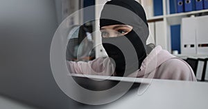 Swindler in balaclava looks through magnifying glass at computer monitor