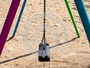 Swing on sandy children`s playground with black plastic seat hanging on chains.