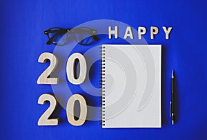 Symbol Happy new year 2020 year, with plywood number 2020, notebook, black vintage glasses and a pencil on blue paper background