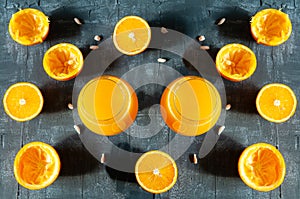 Symmetrically positioned brandy glasses with fresh orange juice. Next to the glass are sliced oranges and zest. Dark background.