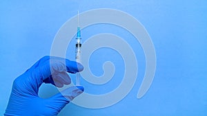 A syringe containing the coronavirus vaccine is held by a blue-gloved hand on a blue background in close-up.