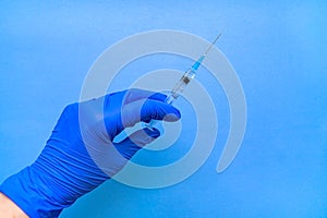 A syringe containing the coronavirus vaccine is held by a blue-gloved hand on a blue background in close-up.