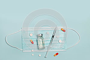 Syringe with medicine from glass vial , medical mask and color pills on light blue background, flat lay, copyspace