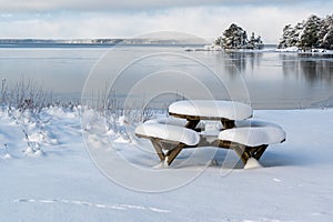 Table with benches covered in snow infront of lake