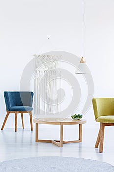 Table between blue and yellow armchair in white living room interior with lamp. Real photo