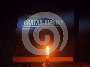 On the table is a book on which the inscription "Holy Bible" in Russian is illuminated. A candle is burning next to it