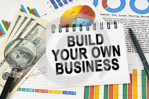 On the table are business charts, dollars and a notepad with the inscription - Build Your Own Business