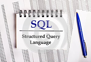 On the table are charts and reports, on which lie a blue pen and a notebook with the word SQL Structured Query Language