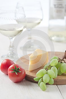 On the table is cheese, tomates, grapes, olive oil, parsley, two wineglasses and a bottle of wine.