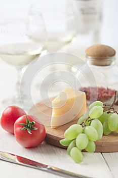 On the table is cheese, tomates, grapes, olive oil, parsley, two wineglasses and a bottle of wine.