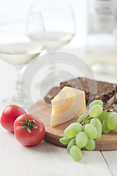 On the table is cheese, tomates, grapes, olive oil, parsley, two wineglasses and a bottle of wine.