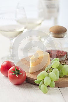 On the table is cheese, tomates, grapes, olive oil, parsley, two wineglasses and a bottle of wine.