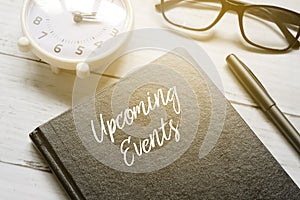 Table clock,eyeglasses,pen and notebook written with UPCOMING EVENTS on white wooden background with sun flare