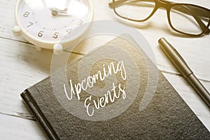 Table clock,eyeglasses,pen and notebook written with UPCOMING EVENTS on white wooden background with sun flare photo