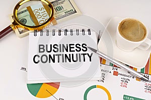 On the table is a cup of coffee, money, business charts and a notepad with the inscription - Business Continuity