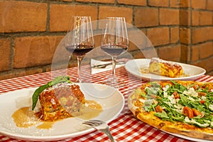 Table in the dining room of an Italian restaurant with pizza to share and individual plates of lasagna and two glasses of red wine