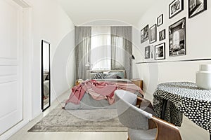 Table with doted tablecloth in bright bedroom interior with white empty wall, mirror and king size bed with grey bedding and