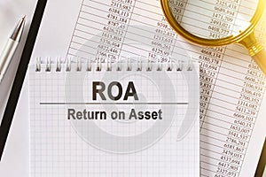 On the table are financial reports, a pen, a magnifying glass and a notebook with the inscription - ROA