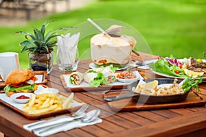 Table full of fresh food - burger with fries, pan with seafood, chicken wraps and young coconut