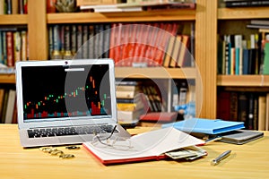On the table golden bitcoins, notebook, eyeglases and laptop with stock exchange graph on screen, blurred shelf with books in the
