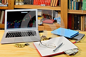 On the table golden bitcoins, notebook, eyeglases and laptop with stock exchange graph on screen, blurred shelf with books in the