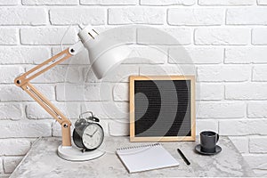 Table lamp, empty blank letter board, notebook and alarm clock on the table, against the background of a white brick wall.