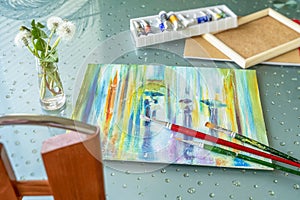 Table with a lying oil painting on canvas, box of paints in tubes, brushes and bouquet of fluffy dandelions. Romantic