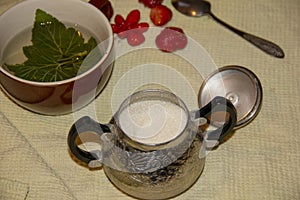 On the table is a Mug, sugar bowl, spoon and strawberry. In a mug lies a piece of currant and poured tea. tea time