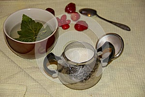 On the table is a Mug, sugar bowl, spoon and strawberry. In a mug lies a piece of currant and poured tea. tea time