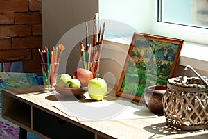 Table with paint tools, picture and tasty apples in artist's workshop