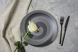 Table place setting with white rose, black plate and cutlery on textured gray background. Top view, flat lay