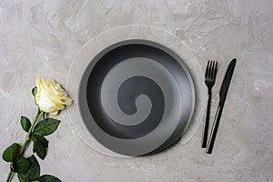 Table place setting with white rose, black plate and cutlery on textured gray background. Top view, flat lay
