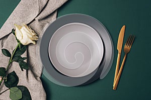 Table place setting with white rose flower, plates and golden cutlery on dark green background. Top view, flat lay