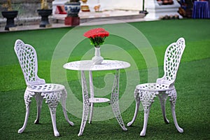 Table with red rose flowers in vase and blank two chair on green grass floor background