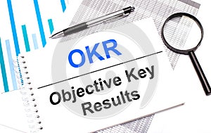 On the table are reports, diagrams, a pen, a magnifying glass and a white notepad with OKR Objective Key Results. Business concept