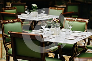 Table served with luxury glasses, cutlery, flowers, green chairs around in cozy restaurant. Nobody in shot. Decorated table for