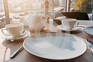 Table setting for breakfast, lunch in sun light. Dinner plate setting with empty plates, cups, teapot near window in