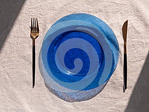 Table setting empty blue glass plate with fork knife on linen cloth top view daylight harsh shadows. Festive dish place