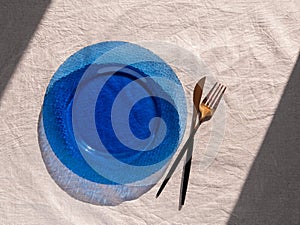 Table setting empty blue glass plate with fork knife on linen cloth top view daylight harsh shadows. Festive dish place