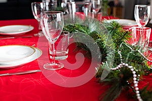 Table setup lunch in red for celebration. Shiny glass cup, red tablecloth, candles and decorations with green Christmas tree.