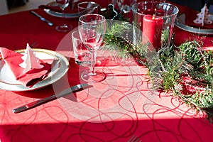 Table setup lunch in red for celebration. Shiny glass cup, red tablecloth, candles and decorations with green Christmas tree.