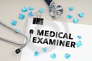 On the table is a stethoscope, a pen, blue crumpled pieces of paper and a sign with the inscription -MEDICAL EXAMINER