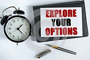 On the table there is a clock, a pen, a notebook and a card on which the text is written - EXPLORE YOUR OPTIONS