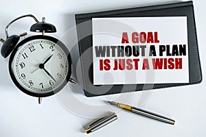 On the table there is a clock, a pen, a notebook and a card on which the text is written - A GOAL WITHOUT A PLAN IS JUST A WISH