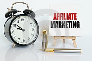 On the table there is a clock, a pen and a stand with a card on which the text is written - AFFILIATE MARKETING