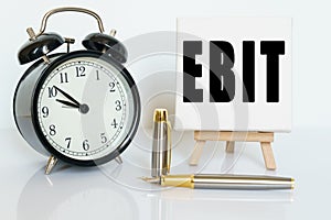 On the table there is a clock, a pen and a stand with a card on which the text is written EBIT. EARNINGS BEFORE INTEREST AND TAXES