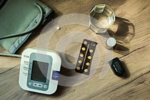 On the table there are pills, a blood pressure measuring device, a glass of water and an oxyometer. An elderly person`s kit. Top